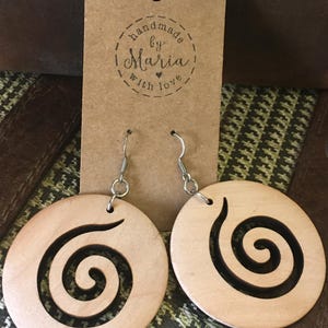 May include: A pair of wooden earrings with a spiral design. The earrings are round and light-colored, with a dark spiral cut out of the wood. The earrings hang from silver-colored hooks. A brown tag behind the earrings reads "handmade by Maria with love."
