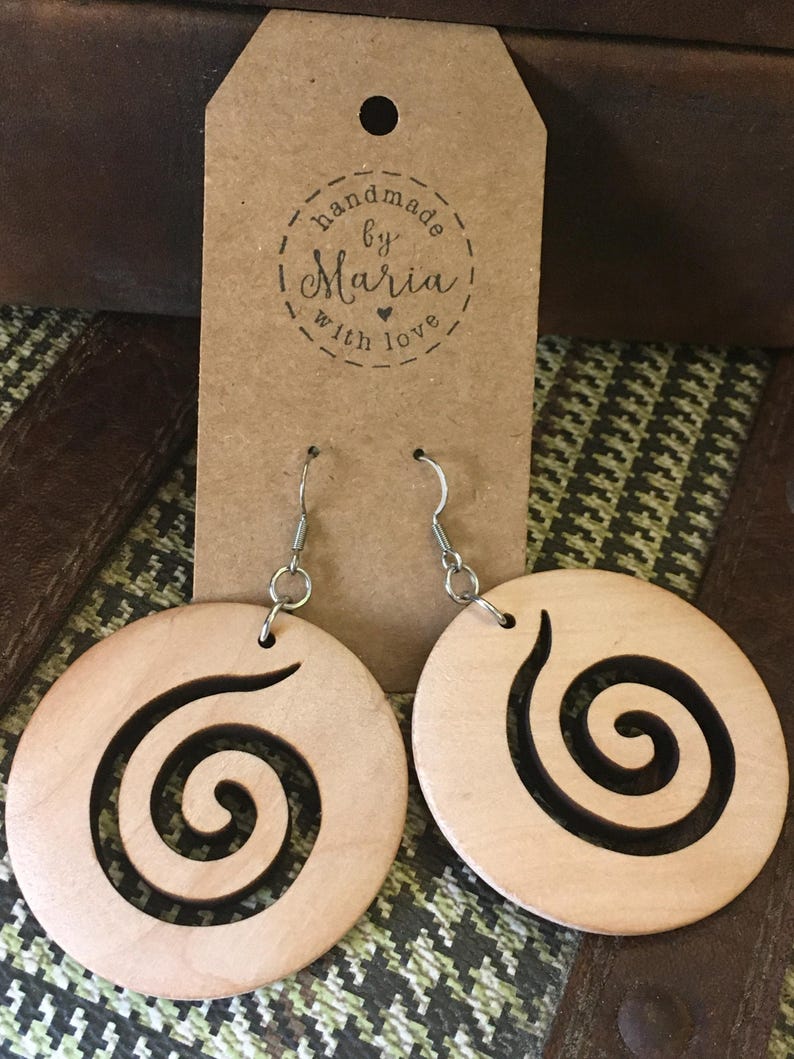 May include: A pair of wooden earrings with a spiral design. The earrings are round and light-colored, with a dark spiral cut out of the wood. The earrings hang from silver-colored hooks. A brown tag behind the earrings reads "handmade by Maria with love."