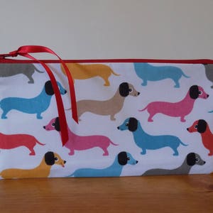 May include: A white zippered pouch with a red zipper pull and red trim. The pouch is decorated with a repeating pattern of colorful dachshund dogs.