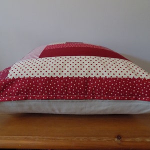 Puede incluir: Coj&iacute;n de patchwork rojo y blanco con un respaldo de color crema. El coj&iacute;n est&aacute; hecho de diferentes telas con varios patrones, incluyendo lunares y estrellas.