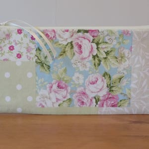 May include: A patchwork fabric zippered pouch with a floral design in shades of pink, green, and blue. The pouch has a white zipper and a white ribbon.