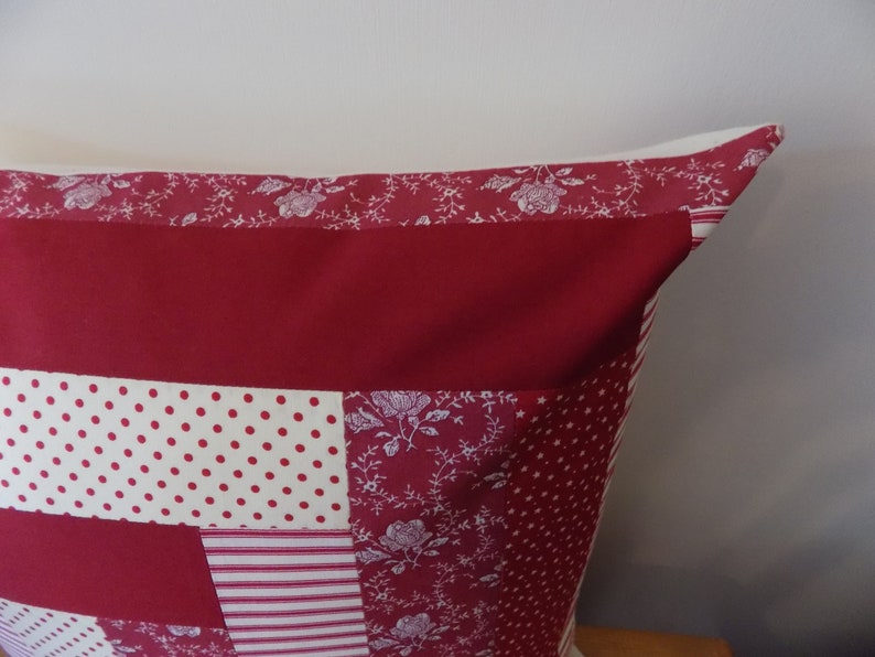 Puede incluir: Una almohada de patchwork rojo, blanco y crema con un patr&oacute;n floral. La almohada est&aacute; hecha de diferentes telas y tiene una forma cuadrada.