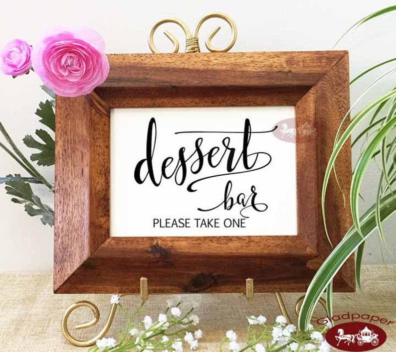 May include: A wooden framed sign with the words "dessert bar" in elegant black script. The sign also includes the text "PLEASE TAKE ONE". The frame is supported by a gold stand and decorated with pink flowers.