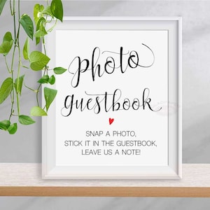 May include: A framed white sign with the words "photo guestbook" in elegant black script, a small red heart, and instructions to snap a photo, stick it in the guestbook, and leave a note. Green leaves hang from the top left.