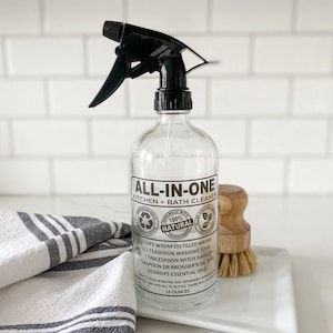 May include: A clear glass spray bottle with a black trigger nozzle. The label reads ALL-IN-ONE KITCHEN + BATH CLEANER and lists ingredients including 3 cups warm distilled water, 1/2 teaspoon washing soda, 1 tablespoon witch hazel, and 20 drops essential oils.