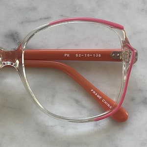 May include: Clear plastic eyeglasses with pink accents. The arms are orange and the frame says "PK 52-16-135" and "FRAME CHINA".