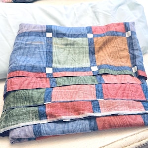 May include: A folded fabric sheet featuring a multi-colored plaid pattern with squares in shades of blue, green, red, and orange, separated by dark blue grid lines and small white accents. A solid light blue fabric piece is tucked behind the plaid sheet.