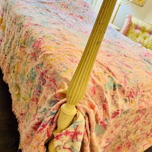 May include: A textured bedspread with a multi-colored tie-dye pattern featuring shades of pink, blue, and yellow, draped over a bed with a cream-colored wooden post. The fabric has a frayed, fringed edge and a crinkled, uneven surface texture.