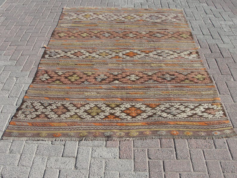 K&ouml;nnte beinhalten: Ein rechteckiger, handgewebter Teppich mit geometrischem Muster. Der Teppich hat horizontale Streifen in Braun-, Orange- und Beiget&ouml;nen mit rautenf&ouml;rmigen Motiven. Der Teppich hat einen Fransenrand und liegt auf einer Ziegeloberfl&auml;che.