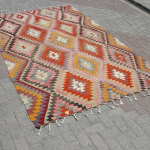 Op de afbeelding: Een kleurrijk, handgeweven tapijt met een geometrisch ruitpatroon in tinten rood, oranje, geel, roze en grijs. Het tapijt heeft een franje rand.