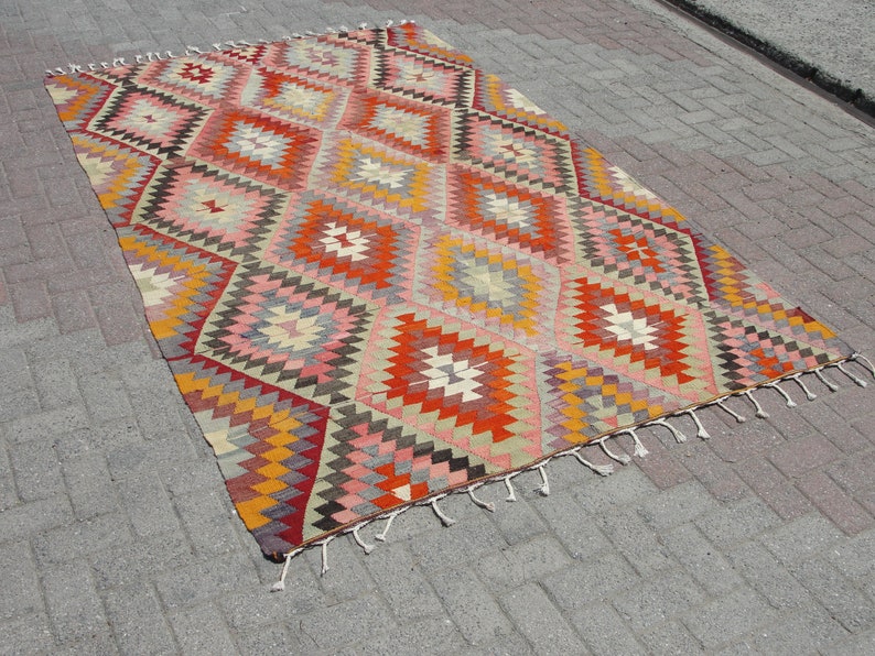 Op de afbeelding: Een kleurrijk, handgeweven tapijt met een geometrisch ruitpatroon in tinten rood, oranje, geel, roze en grijs. Het tapijt heeft een franje rand.