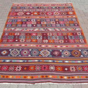 May include: A rectangular, hand-woven rug with a vibrant, geometric pattern. The rug features horizontal stripes in shades of red, orange, purple, and pink, with detailed geometric shapes and fringe along the edges. The rug is suitable for home decor.