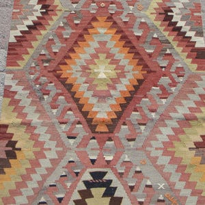 May include: A rectangular, hand-woven rug with a geometric pattern. The rug features a central diamond design in shades of orange, red, and brown, surrounded by repeating zigzag patterns in gray, pink, and yellow. The rug's dimensions are not visible.
