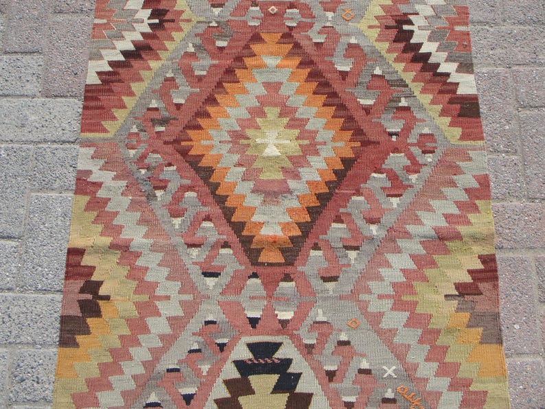 May include: A rectangular, hand-woven rug with a geometric pattern. The rug features a central diamond design in shades of orange, red, and brown, surrounded by repeating zigzag patterns in gray, pink, and yellow. The rug's dimensions are not visible.