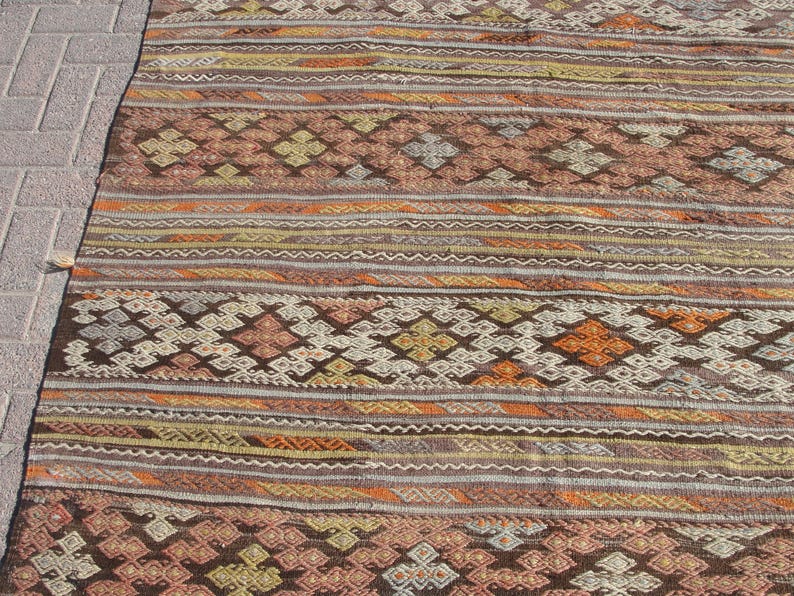 K&ouml;nnte beinhalten: Ein rechteckiger Teppich mit geometrischem Muster in Braun-, Orange- und Cremet&ouml;nen. Das Design des Teppichs umfasst Rauten- und Streifenmotive. Er besteht aus gewebtem Material, wodurch er sich strukturiert anf&uuml;hlt.