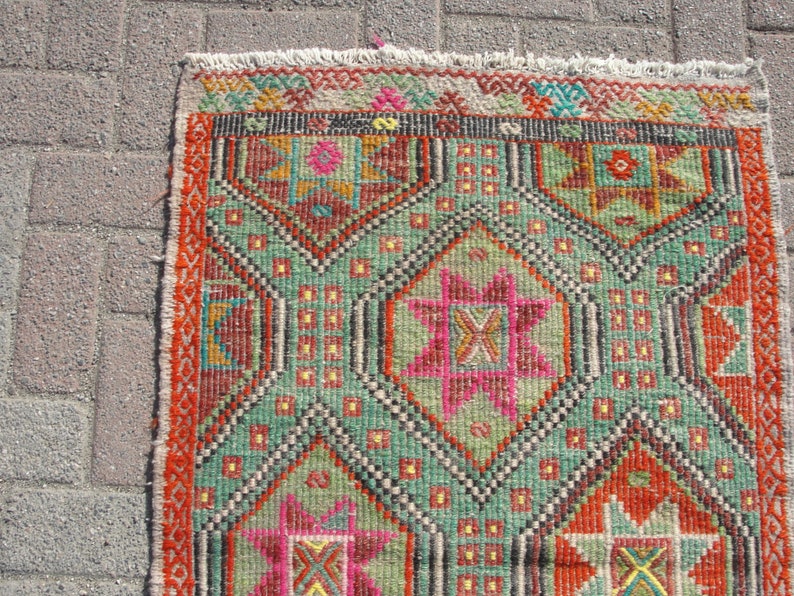Puede incluir: Una alfombra colorida con un patr&oacute;n geom&eacute;trico en tonos rojos, verdes, rosas y amarillos. La alfombra tiene un borde de flecos.