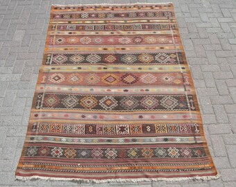 May include: A vintage Turkish rug with a colorful geometric pattern in shades of red, orange, brown, and beige. The rug has a fringed edge and is made of wool.