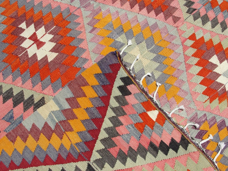 Op de afbeelding: Een kleurrijk tapijt met een geometrisch patroon in tinten rood, oranje, roze, grijs en wit. Het tapijt heeft een franje rand.