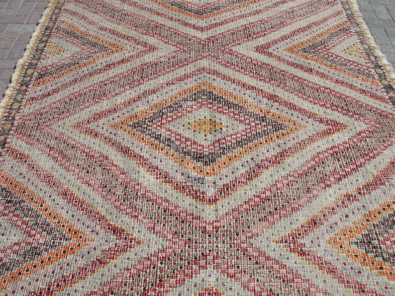 May include: A close-up of a beige, red, orange, and black patterned rug with a diamond pattern. The rug has a woven texture.
