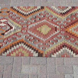 May include: A rectangular, patterned rug with a geometric design. The rug features a repeating pattern of diamonds and zigzags in shades of red, orange, gray, and beige. The rug has a fringed edge.