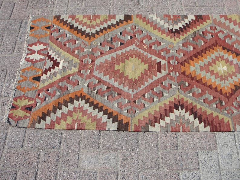 May include: A rectangular, patterned rug with a geometric design. The rug features a repeating pattern of diamonds and zigzags in shades of red, orange, gray, and beige. The rug has a fringed edge.