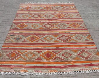May include: A rectangular, hand-woven rug with a geometric pattern. The rug features diamond and stripe designs in shades of red, orange, and beige. The edges are finished with a fringe. This rug is suitable for home decor.