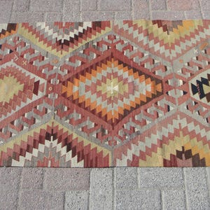 May include: A rectangular rug with a geometric pattern in shades of red, orange, yellow, and gray. The design features diamond and zigzag shapes, creating a textured appearance. The rug is placed on a brick surface.