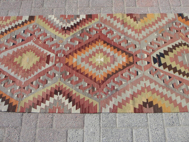 May include: A rectangular rug with a geometric pattern in shades of red, orange, yellow, and gray. The design features diamond and zigzag shapes, creating a textured appearance. The rug is placed on a brick surface.