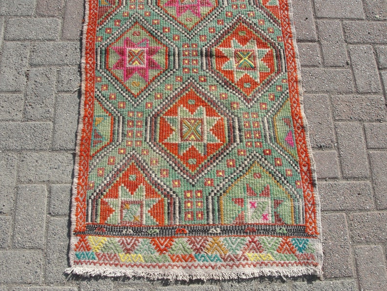 Puede incluir: Una alfombra kilim turca tradicional con un patr&oacute;n geom&eacute;trico en tonos rojos, verdes, naranjas y amarillos. La alfombra tiene un borde de flecos y est&aacute; hecha de lana.