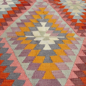 Op de afbeelding: Een kleurrijk tapijt met een geometrisch patroon in tinten oranje, roze, grijs en wit. Het patroon bestaat uit herhalende driehoeken en ruiten.