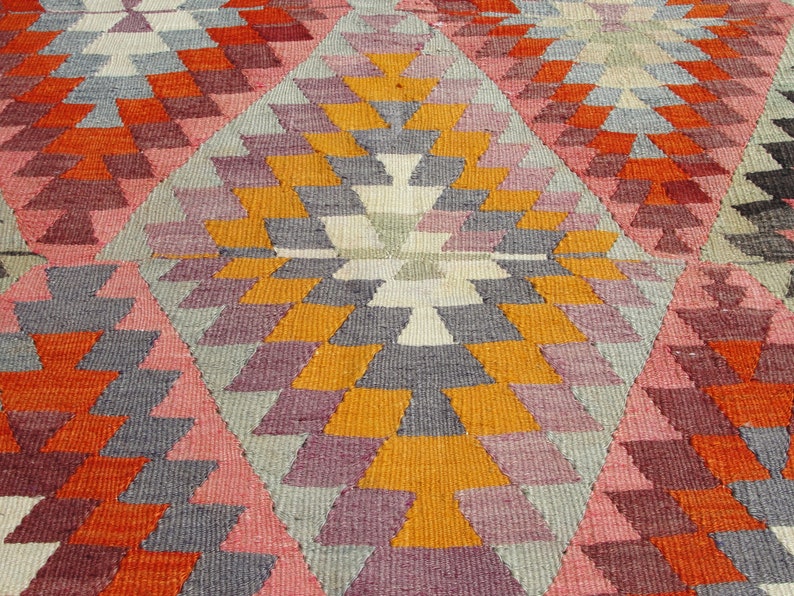 Op de afbeelding: Een kleurrijk tapijt met een geometrisch patroon in tinten oranje, roze, grijs en wit. Het patroon bestaat uit herhalende driehoeken en ruiten.