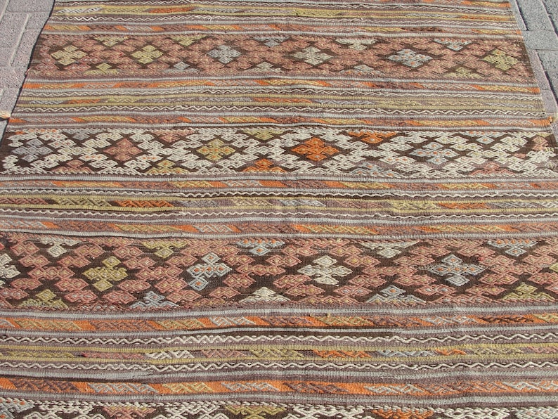 K&ouml;nnte beinhalten: Ein rechteckiger, gewebter Teppich mit geometrischem Muster. Der Teppich weist horizontale Streifen in Braun-, Orange- und Cremet&ouml;nen mit rauten- und kreuzartigen Motiven auf. Der Teppich ist wahrscheinlich aus Naturfasern gefertigt.