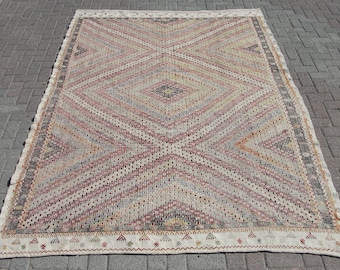 May include: Rectangular woven rug with a geometric diamond pattern in shades of cream, red, gray, and black. The rug has a cream-colored border with small decorative elements. The rug is placed on a brick surface.
