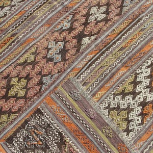 K&ouml;nnte beinhalten: Nahaufnahme eines gewebten Teppichs mit geometrischem Muster. Der Teppich weist verschiedene Farben auf, darunter Braun, Orange und Creme. Das Design umfasst Rauten- und Zickzackmuster. Der Teppich hat eine strukturierte Oberfl&auml;che und einen Fransenrand.