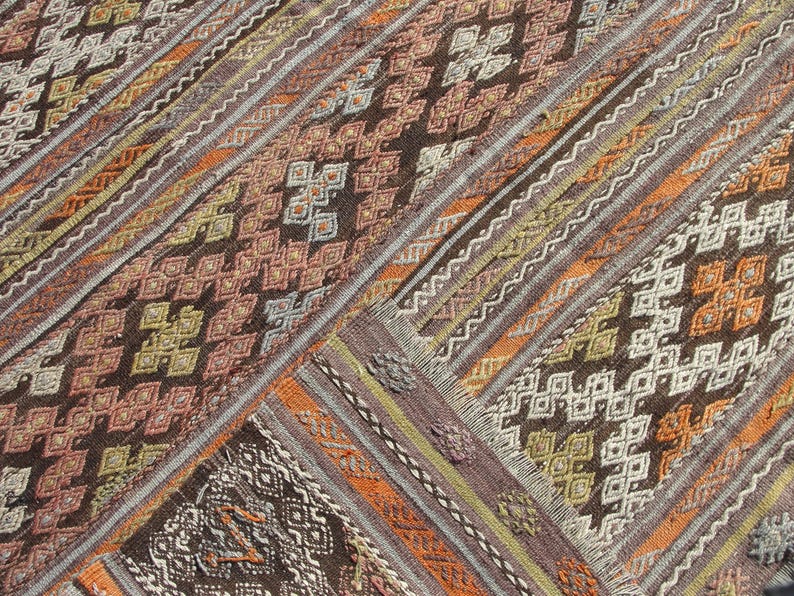 K&ouml;nnte beinhalten: Nahaufnahme eines gewebten Teppichs mit geometrischem Muster. Der Teppich weist verschiedene Farben auf, darunter Braun, Orange und Creme. Das Design umfasst Rauten- und Zickzackmuster. Der Teppich hat eine strukturierte Oberfl&auml;che und einen Fransenrand.