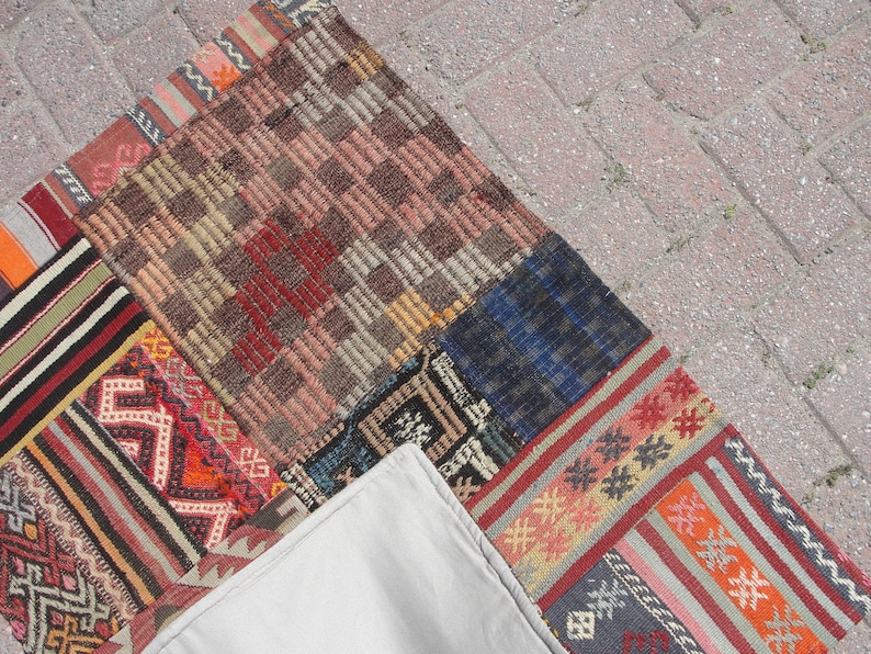 May include: A patchwork rug made of different colored fabrics with geometric patterns. The rug is made up of several smaller rugs sewn together. The rug is on a gray concrete surface.