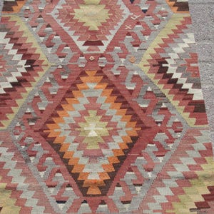 May include: A rectangular, patterned rug with a geometric design. The rug features a diamond pattern in shades of red, orange, gray, and beige. The rug is made of woven material and is suitable for use in a living room or bedroom.