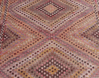 May include: A close-up of a red, brown, and white patterned rug with a diamond-shaped design. The rug has a woven texture and is made of wool.