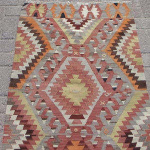 May include: A rectangular, hand-woven rug with a geometric pattern. The rug features a central diamond design in shades of red, orange, and yellow, surrounded by zig-zag and triangular patterns in brown, gray, and white. The rug has a fringed edge.
