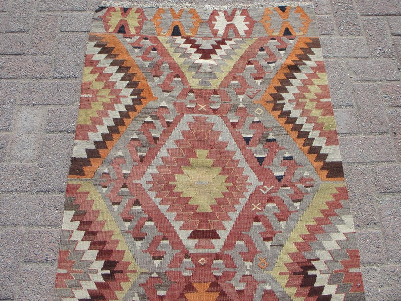 May include: A rectangular, hand-woven rug with a geometric pattern. The rug features a central diamond design in shades of red, orange, and yellow, surrounded by zig-zag and triangular patterns in brown, gray, and white. The rug has a fringed edge.