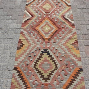 May include: A rectangular, patterned rug with a geometric design. The rug features a repeating diamond pattern in shades of orange, brown, beige, and gray. The rug is placed on a brick surface.