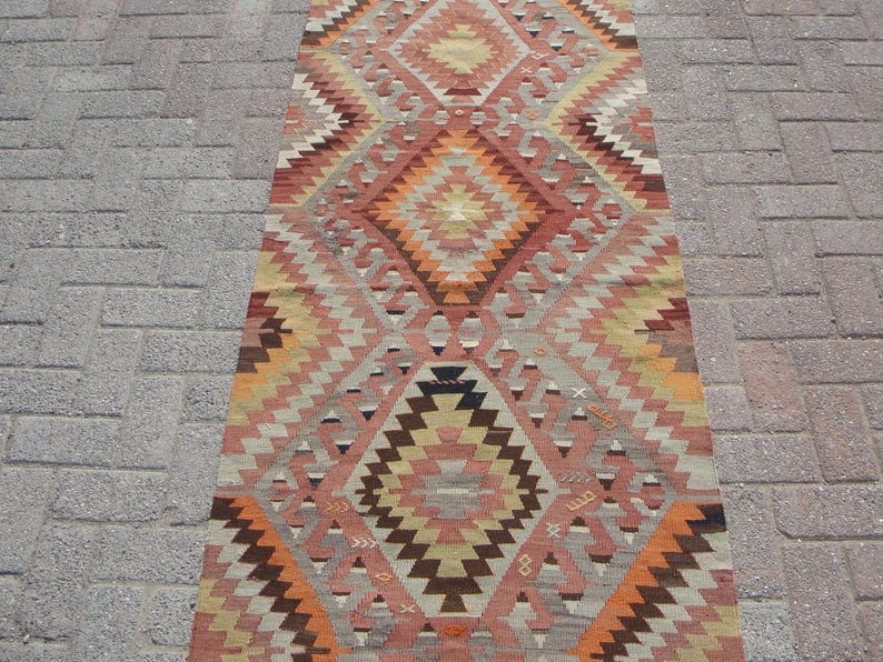 May include: A rectangular, patterned rug with a geometric design. The rug features a repeating diamond pattern in shades of orange, brown, beige, and gray. The rug is placed on a brick surface.