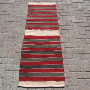 May include: A long, narrow, striped rug with alternating bands of red, brown, and cream. The rug is woven with a simple, traditional design.