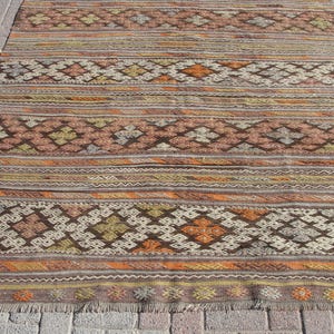 K&ouml;nnte beinhalten: Ein rechteckiger Teppich mit einem Muster in warmen Farben wie Braun, Orange und Creme. Der Teppich hat geometrische Designs und horizontale Streifen mit einem Fransenrand. Er liegt auf einer Ziegeloberfl&auml;che.