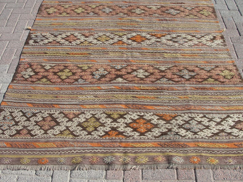 K&ouml;nnte beinhalten: Ein rechteckiger Teppich mit einem Muster in warmen Farben wie Braun, Orange und Creme. Der Teppich hat geometrische Designs und horizontale Streifen mit einem Fransenrand. Er liegt auf einer Ziegeloberfl&auml;che.