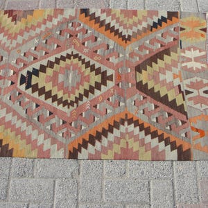 May include: A flat-weave rug with a geometric pattern in shades of orange, pink, brown, and beige. The rug features diamond and zigzag designs, with a gray border and fringe. This rug is suitable for home decor.