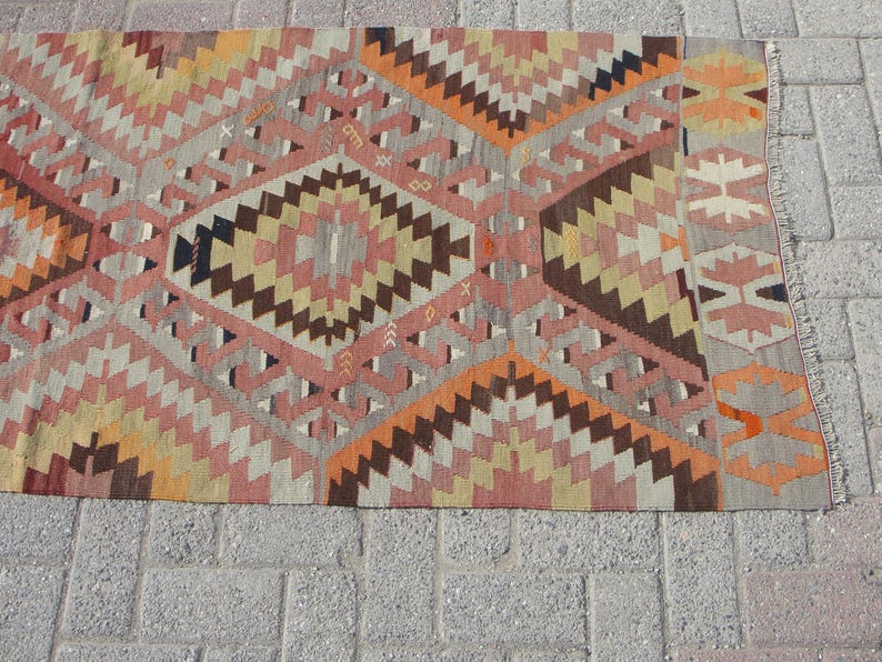 May include: A flat-weave rug with a geometric pattern in shades of orange, pink, brown, and beige. The rug features diamond and zigzag designs, with a gray border and fringe. This rug is suitable for home decor.