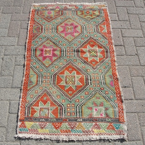 Puede incluir: Una alfombra turca vintage con un patr&oacute;n geom&eacute;trico en tonos rojos, verdes, naranjas y rosas. La alfombra tiene un borde con flecos y est&aacute; hecha de lana.