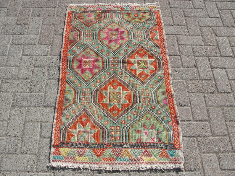 Puede incluir: Una alfombra turca vintage con un patr&oacute;n geom&eacute;trico en tonos rojos, verdes, naranjas y rosas. La alfombra tiene un borde con flecos y est&aacute; hecha de lana.