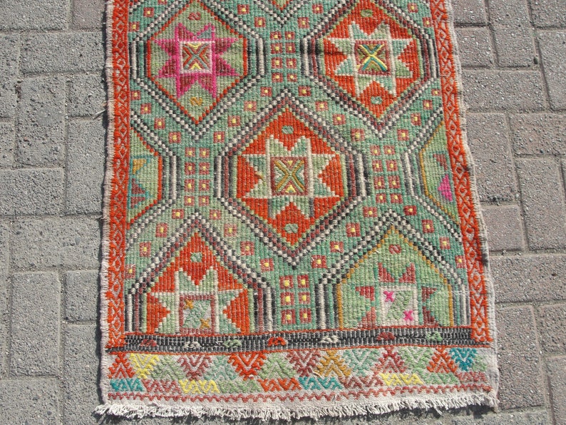 Puede incluir: Una alfombra kilim turca tradicional con un patr&oacute;n geom&eacute;trico en tonos rojos, verdes, naranjas y amarillos. La alfombra tiene un borde con flecos y est&aacute; hecha de lana.